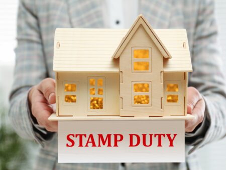 A person carrying a wooden model house with a sign that says 'stamp duty'