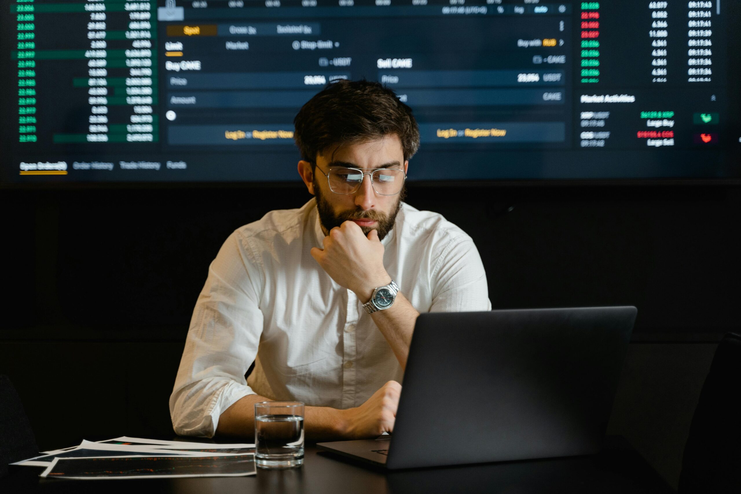 An image of a man sitting by a laptop with financial data behind im on a screen